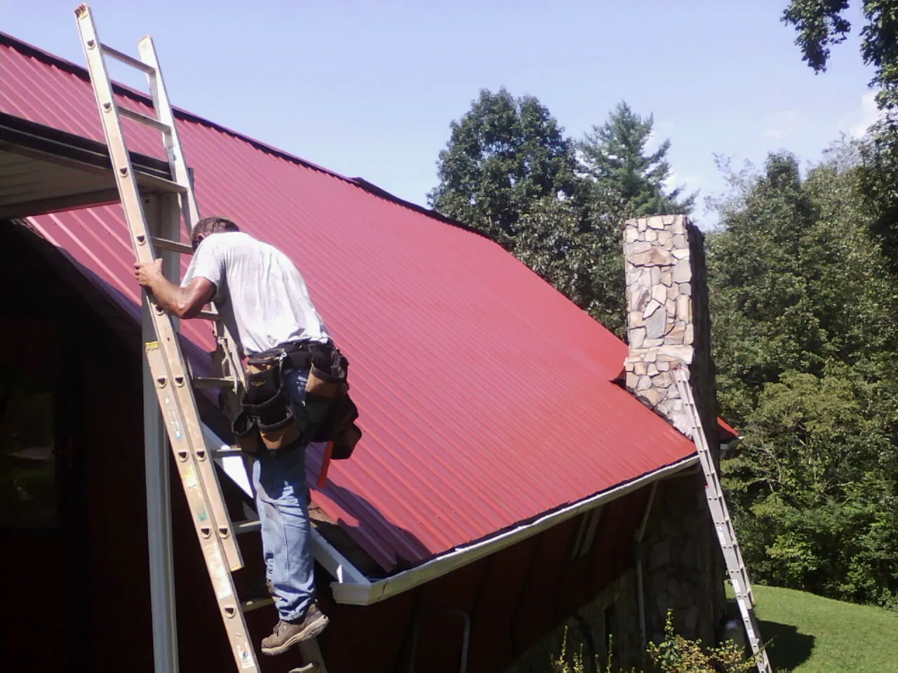 Roofer on a ladder beside a red metal roof and stone chimney — Soffit & Fascia Repair by Roofing Brothers in Elkin